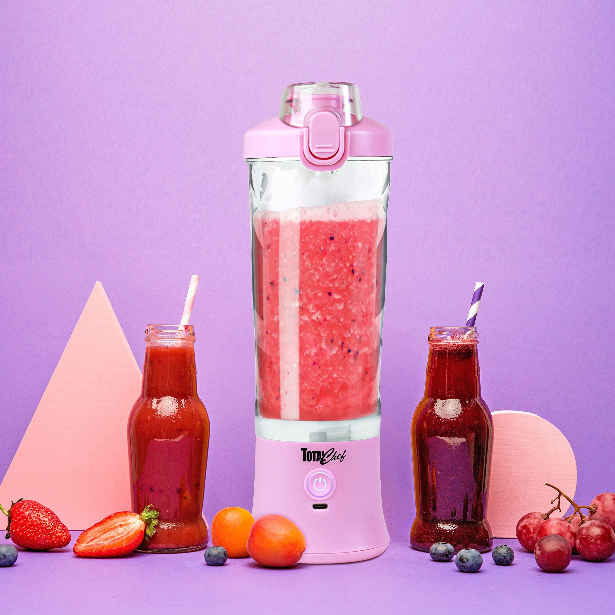 Lifestyle image of the Total Chef USB chargeable blender filled with bright red smoothie on a purple background with grapes, tangerines, strawberries, blueberries, and two glass bottles with straws filled with red and purple smoothies arranged around