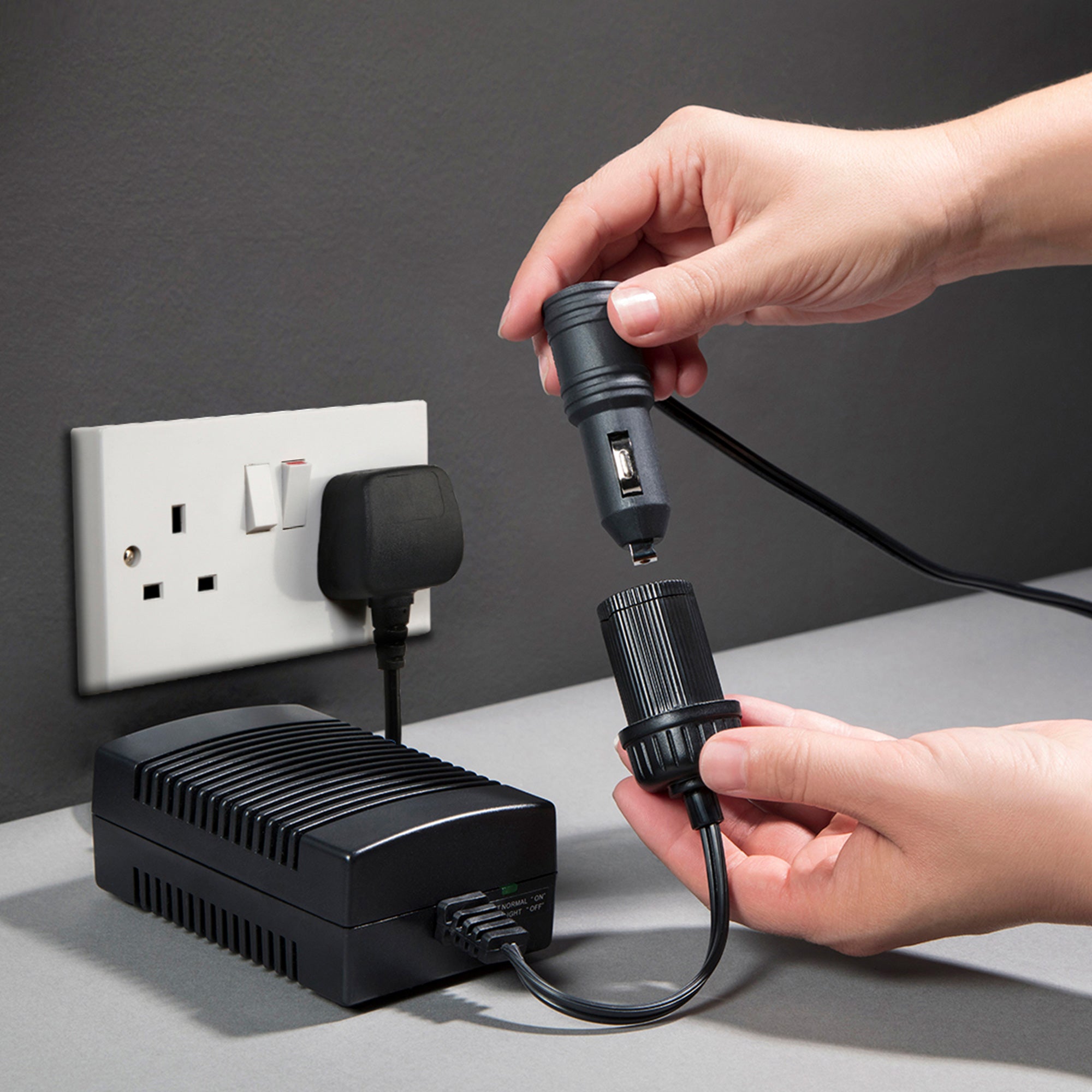 Koolatron AC to DC power adapter plugged into a white indoor AC receptacle on a dark gray wall with a person’s hands inserting a 12V plug into the DC adapter end