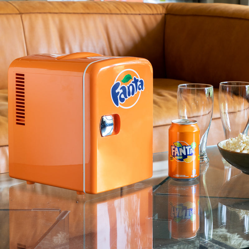 Lifestyle image of Coca-Cola Fanta 6 can mini fridge, partly open with cans inside, on a glass coffee table with a tan leather sofa behind it and a can of Fanta and an empty glass to the right