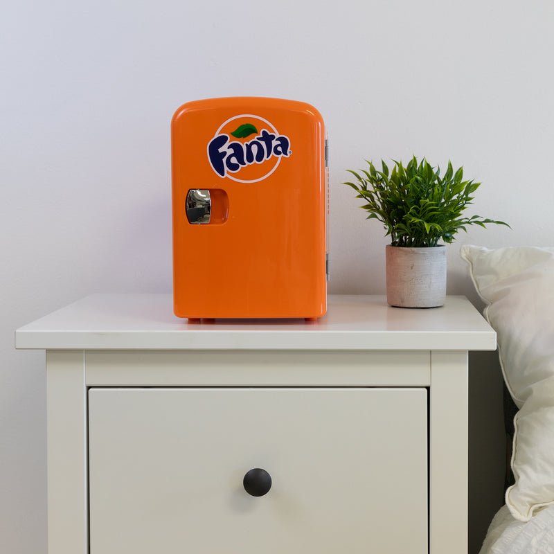 Lifestyle image of Coca-Cola Fanta 6 can mini fridge, closed, on a white bedside table with a plant in a white pot on its right and a white painted wall behind
