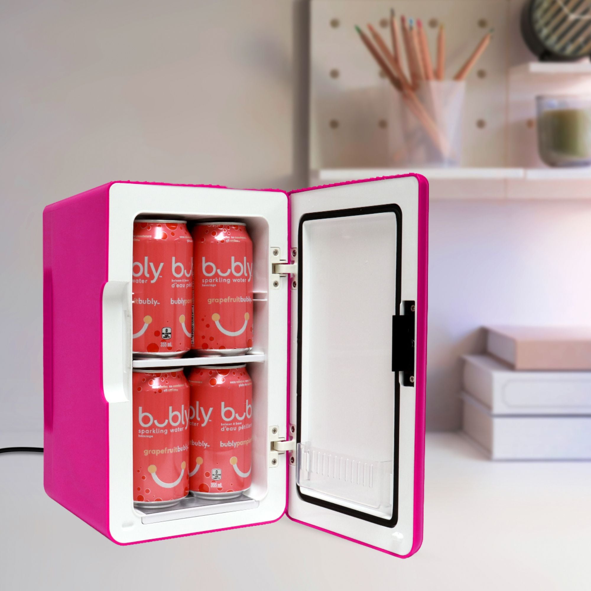 Koolatron 8 can mini fridge, open with 8 soft drink cans inside, on a white desk with a cup of pencils and a pile of books in the background