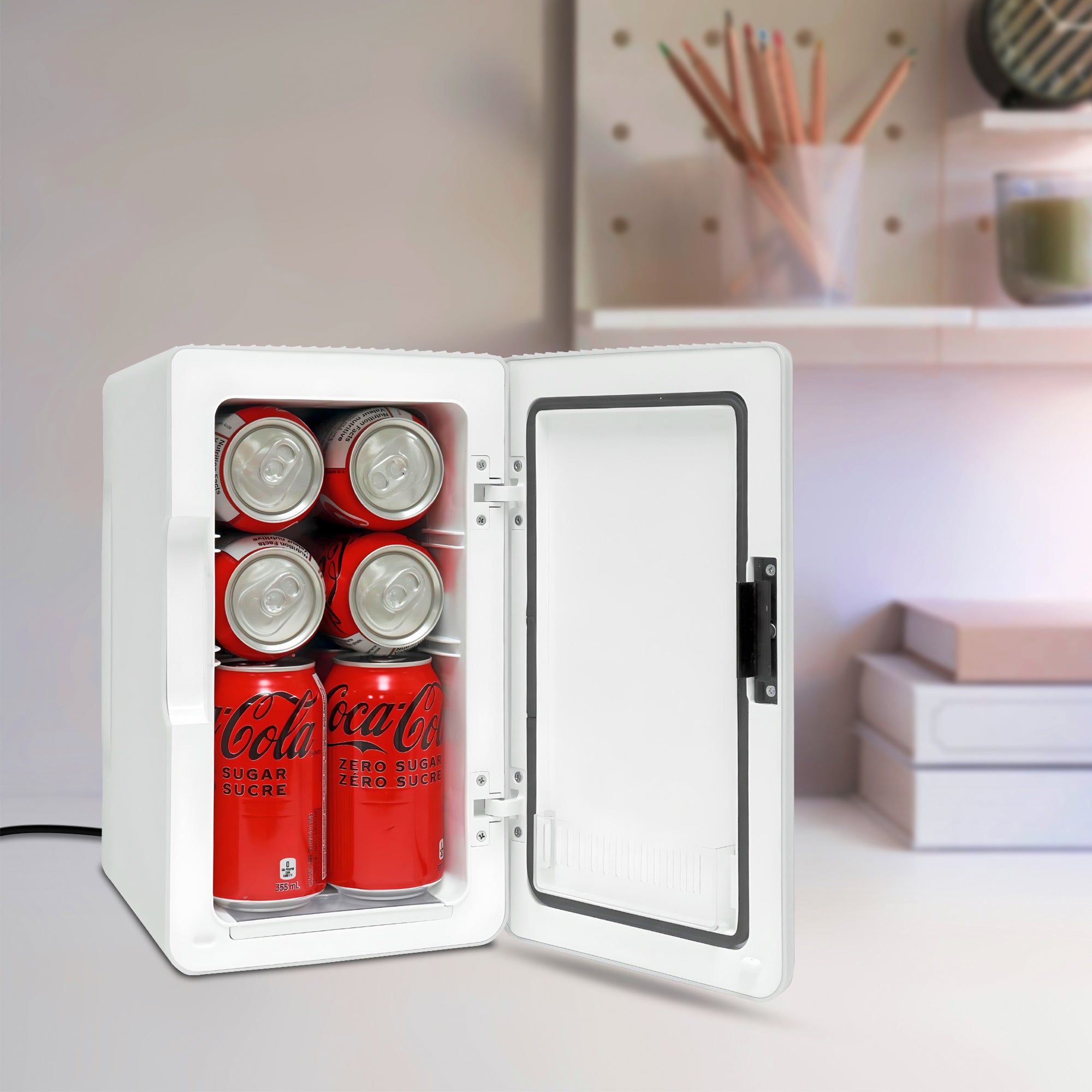 Koolatron 8 can mini fridge, open with 8 soft drink cans inside, on a white desk with a cup of pencils and a pile of books in the background