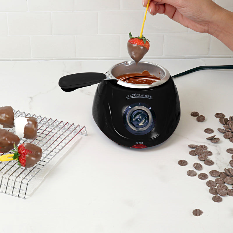 Lifestyle image of a hand dipping a strawberry in melted chocolate in the chocolatiere on a white countertop. There is a cooling rack to the left with dipped strawberries and marshmallows on it and unmelted chocolate discs scattered on the counter to the right