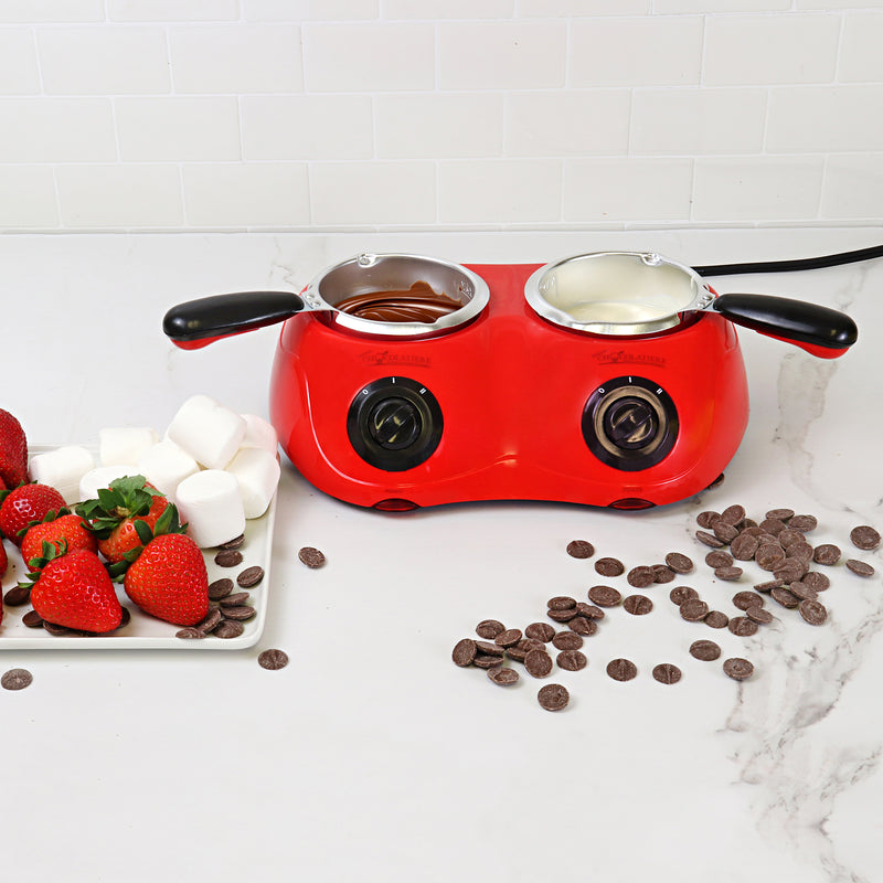 Lifestyle image of the chocolatiere on a white marbled countertop with melted milk chocolate in one pot and white chocolate in the other. There is a cooling rack to the left with strawberries and marshmallows on it and unmelted chocolate discs scattered on the counter to the right