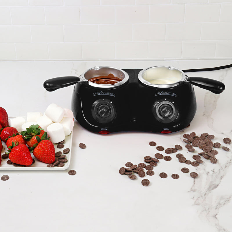 Lifestyle image of the chocolatiere on a white marbled countertop with melted milk chocolate in one pot and white chocolate in the other. There is a plate to the left with strawberries and marshmallows on it and unmelted chocolate discs scattered on the counter to the right