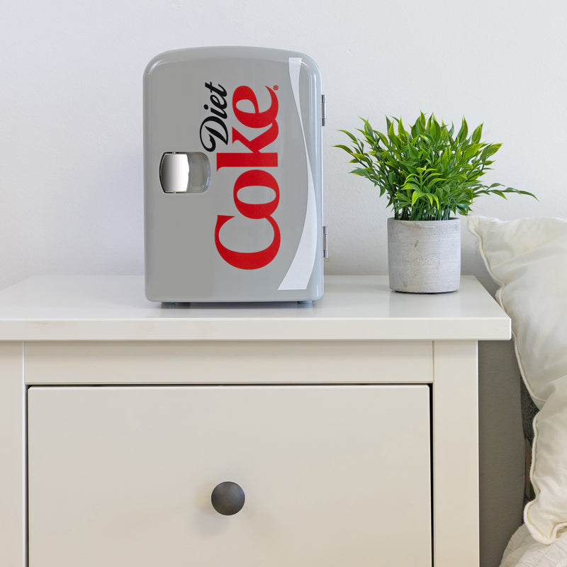 Lifestyle image of Coca-Cola Diet Coke 6 can mini fridge, closed, on a white bedside table with a plant in a white pot on its right and a white painted wall behind