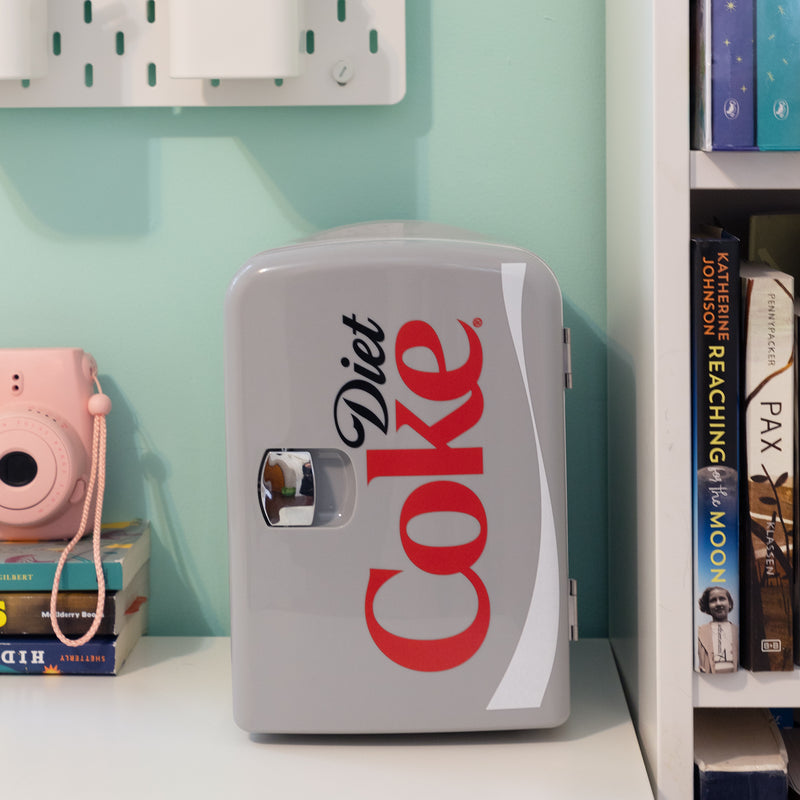 Lifestyle image of Coca-Cola Diet Coke 6 can mini fridge, closed, on a white desktop with an aqua wall behind. There is a stack of books and a pink camera to the left of the fridge and a bookshelf to the right