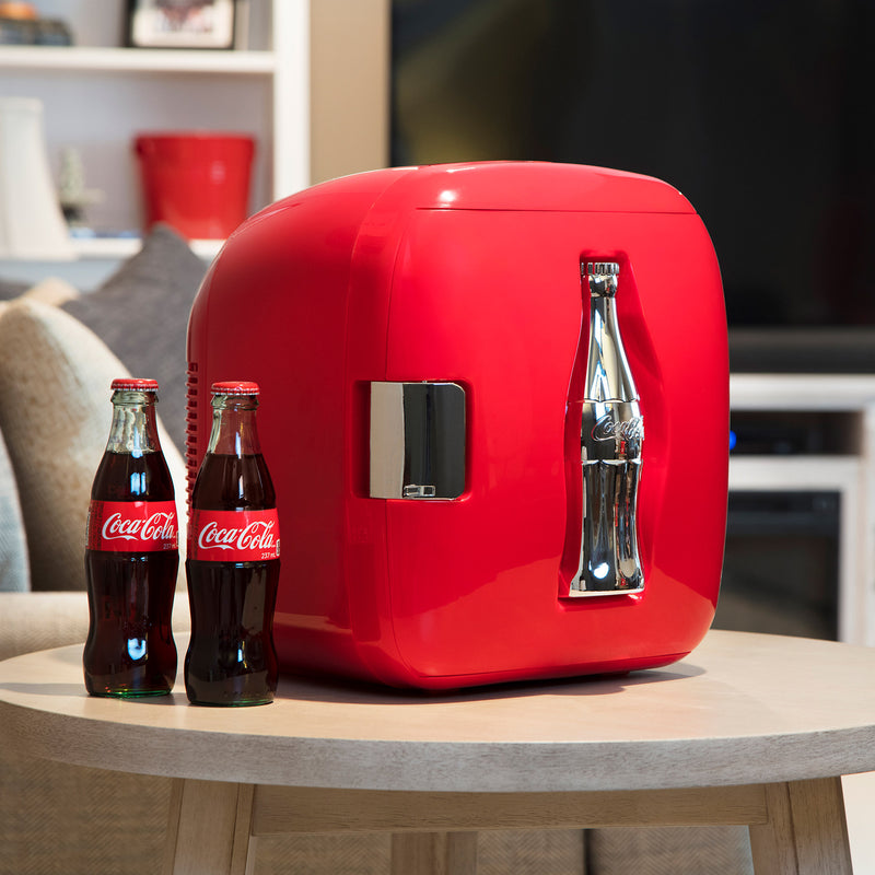 Lifestyle image of Coca-Cola 12 can cooler/warmer, closed, on light beige side table with two glass bottles of Coke beside it and a couch, bookcase, and TV in the background