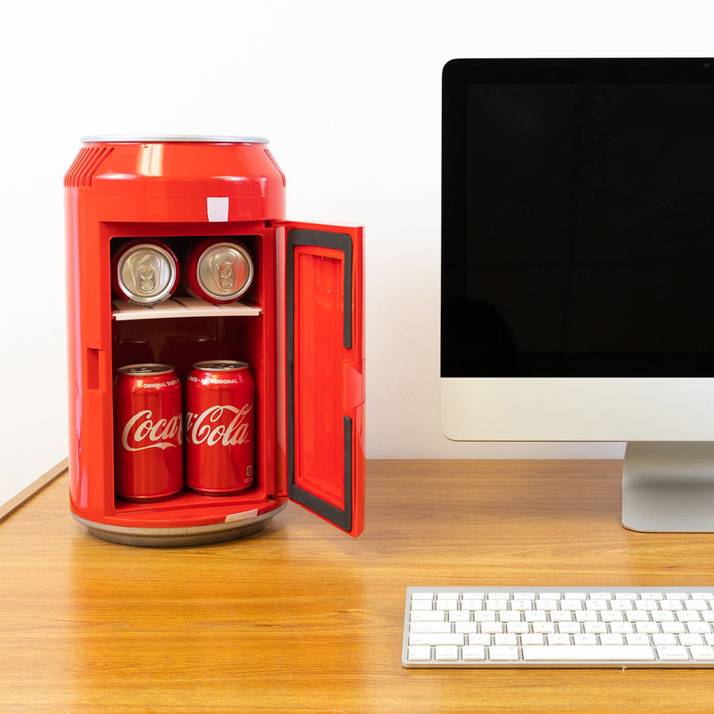 Lifestyle image of Coca-Cola can-shaped mini fridge, open with 6 cans of Coke inside, on a wooden desktop with a computer monitor and keyboard beside it